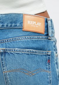 Blugi din denim albastru, cu o etichetă din piele maro deschis inscripționată cu "REPLAY DIN 1981." Designul include cusături clasice la buzunare și un finisaj texturat.