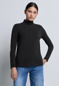 Musta pitkähiha turtleneck-paita, jossa on istuva muotoilu, sileä pinta ja minimaalinen ommel. Yhdistetty sinisiin farkkuihin. Yksinkertainen tausta.