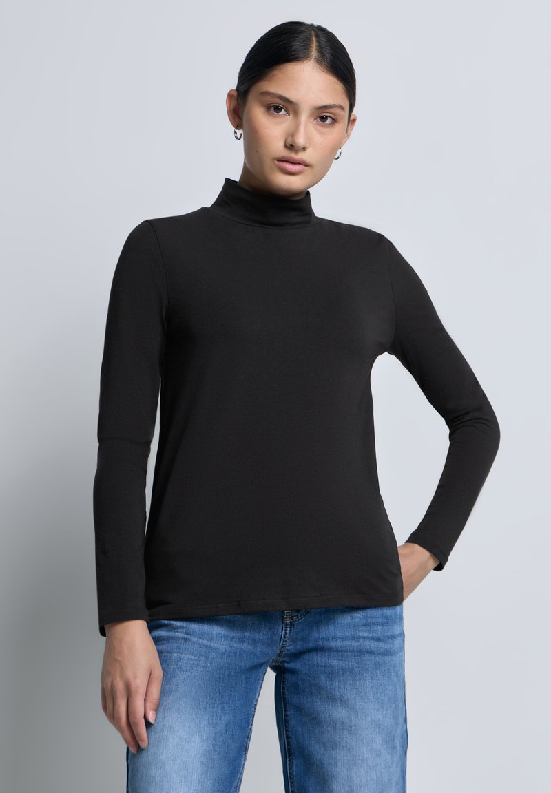 Musta pitkähiha turtleneck-paita, jossa on istuva muotoilu, sileä pinta ja minimaalinen ommel. Yhdistetty sinisiin farkkuihin. Yksinkertainen tausta.