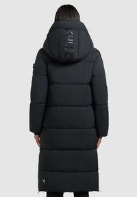 Osoba v dlouhém černém péřovém kabátu s velkou kapucí, na kapuci je viditelné vertikální logo, stojící zády k plochému pozadí.