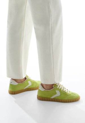 Green suede sneakers with white accents, featuring a textured sole and lace-up front. Leg is clad in cream-colored, slightly tapered pants.
