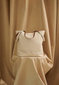 Beige Lederhandtasche mit weicher Textur, ausgestattet mit braunen und schwarzen Trägern sowie zwei silbernen Metallakzenten. Minimalistisches Design ohne sichtbare Logos.