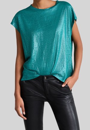 Femme portant un haut scintillant à manches courtes de couleur bleu sarcelle, rentrée dans un pantalon en cuir noir, avec une main dans la poche, sur fond gris.