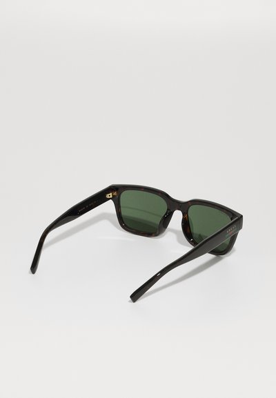 Rechteckige Sonnenbrille mit Schildpatt-Fassung, dunkelgrünen Gläsern und dünnen schwarzen Bügeln, auf einer weißen Oberfläche platziert.