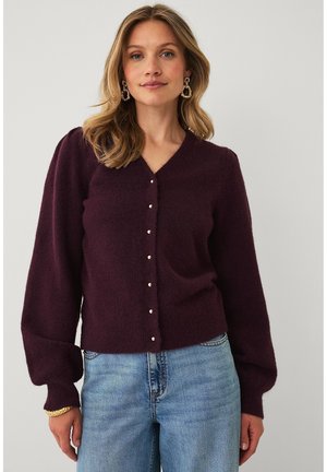 Kvinde med blond hår iført lilla knappet cardigan med puffede ærmer og blå jeans, tilbehør med guldbøjler og armbånd.