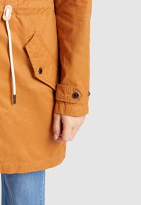 Pomarańczowa, lekka kurtka z regulowanym paskiem w talii, przednią kieszenią i zapinanymi na guziki mankietami. Gładka tekstura i casualowy design.