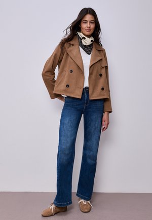 Femme portant une veste courte camel à double boutonnage, un haut blanc, un jean bleu, des chaussures beiges et une écharpe à pois noir et blanc autour du cou.