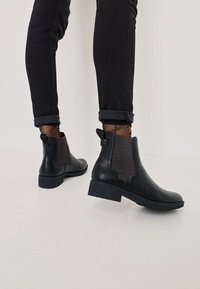 Bocanci negri din piele la gleznă cu panouri elastice laterale, accente texturate gri și toc mic îngrămădit, purtați cu blugi negri cu manșetă.