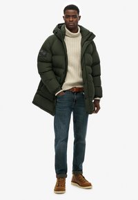 Puchowy zielony kurtka z kapturem, kremowy dziany sweter, niebieskie dżinsy i brązowe buty na sznurówki. Kurtka ma logo na rękawie.