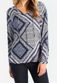 Blouse bleu marine à motifs floraux et géométriques, manches longues, encolure en V et cordons avec accents décoratifs.