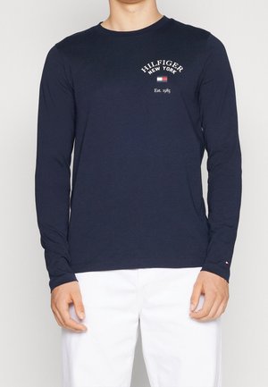 Homme portant une chemise bleu marine à manches longues avec "Hilfiger New York Est. 1985" et un petit logo drapeau sur la poitrine, associée à un pantalon blanc.