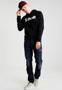Felpa nera con grafica "Schott NYC", jeans di denim blu e sneakers beige. Il modello indossa un cappellino bordeaux e posa con una mano alzata.