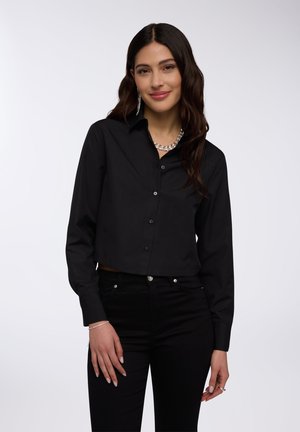 Camicetta nera cropped con bottoni e maniche lunghe, dotata di un colletto e realizzata in tessuto liscio. Abbinata a jeans neri aderenti.