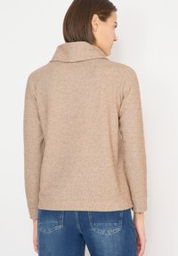 Suéter de punto beige con un cuello ancho, mangas largas y textura acanalada. Se lleva con vaqueros azules, mostrando un ajuste relajado y un diseño casual.