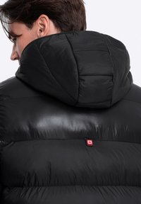 Schwarze Pufferjacke mit gesteppter Kapuze und glatter Textur. Auffälliges rotes Logo nahe dem Kragen, das das praktische Design betont.