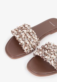 Sandali a scivolo marroni con una striscia decorata con perle e accenti metallici su una superficie testurizzata. Suola piatta con un design semplice e moderno.