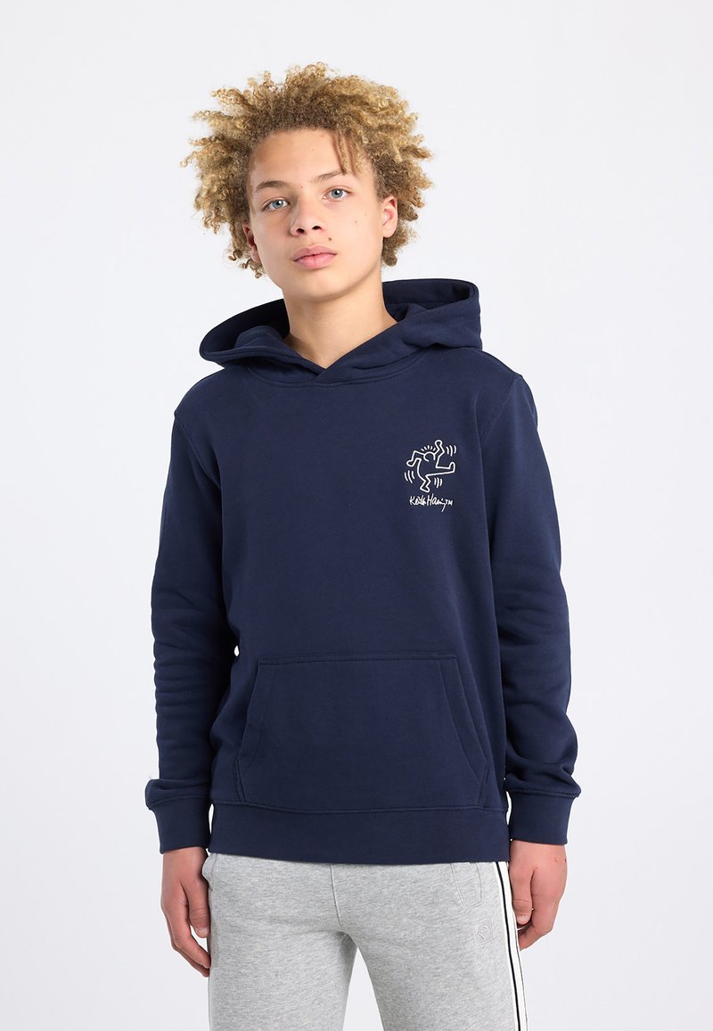 Adolescent aux cheveux blonds bouclés portant un sweat à capuche bleu marine avec un petit motif de Keith Haring et un pantalon de survêtement gris clair avec des bandes latérales.
