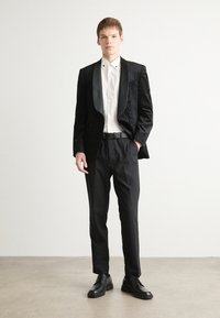 Svart tuxedojacka med texturerat mönster, satinrevers, vit skjorta, svarta byxor och svarta läderskor. Enkel, skräddarsydd design.