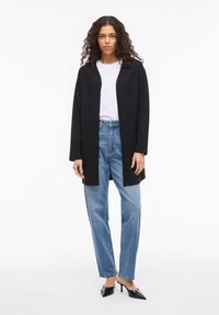 Μαύρο παλτό με ανοιχτό μπροστινό μέρος, μακριά μανίκια, υφασμάτινη υφή, δύο μπροστινές τσέπες· φορεμένο πάνω από λευκό Τ-shirt και συνδυασμένο με ανοιχτόχρωμα μπλε τζιν και μαύρες γόβες.