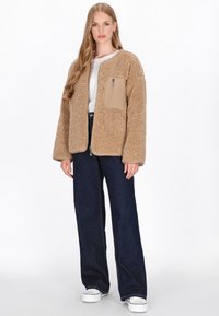 Beige Fleecejacke mit strukturierter Außenseite und einer braunen Reißverschluss-Tasche, kombiniert mit dunkelblauen weit geschnittenen Jeans und weißen Schuhen.