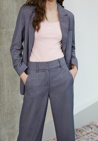 Gray pinstriped suit with relaxed-fit trousers and a blazer. Worn over a light pink tank top with a small white logo. Smooth texture.