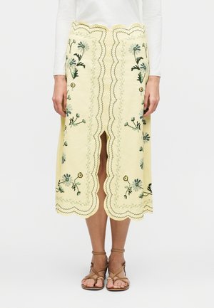 Jasnożółta spódnica do kolan z falistymi brzegami, kwiatowym haftem, przednim rozcięciem, noszona z białym topem i brązowymi sandałami na paski.