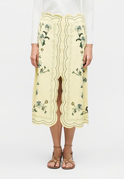 Jupe jaune pâle longueur genou avec bords festonnés, broderie florale, fente devant, portée avec un haut blanc et des sandales marron à brides.