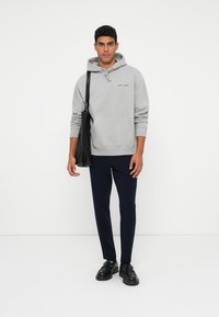 Sweat à capuche gris avec une poche kangourou, détails en lettres noires, associé à un pantalon bleu marine foncé et des chaussures noires. Le modèle tient un sac fourre-tout noir.