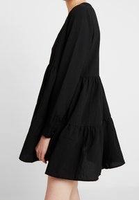 Schwarzes Puffkleid mit langen Ärmeln, gestuftem Design und lockerer Passform. Der Stoff hat eine weiche Textur mit einem subtilen Glanz und Raffung an der Taille.