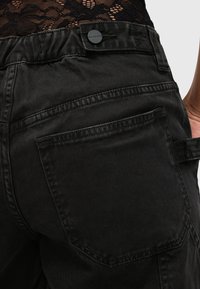 Zwarte katoenen broeken met een rond achterzak, een brede tailleband en een klein stoffen logo-detail nabij de tailleband.