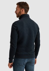 Marineblauer, gesteppter Mantel mit hohem Kragen, aus strukturiertem Stoff, mit langen Ärmeln und einem Reißverschluss am Hals. Sichtbare blaue Jeans.