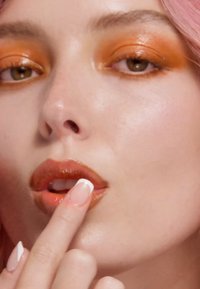 Primo piano di un volto con ombretto arancione e labbra lucide di color marrone. Il dito della modella tocca leggermente il labbro inferiore. Le unghie sono curate con punte bianche.