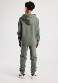 Hellgrüner Baumwoll-Hoodie mit großer Kapuze, kombiniert mit passenden Jogginghosen. Mit gerippten Bündchen und einem lockeren Schnitt, getragen mit weißen Turnschuhen.