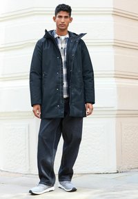 Parka de un navy închis cu glugă și închidere cu nasture tip snap, asortat cu o cămașă în carouri și blugi largi de denim. Adidașii gri completează look-ul.