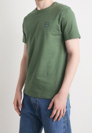 Mann trägt ein schlichtes, olivgrünes T-Shirt mit kurzen Ärmeln und kleinem Logo auf der Brust sowie blaue Jeans. Er steht vor einem schlichten Hintergrund.