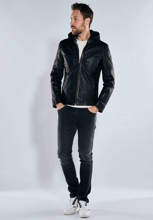 Schwarze Lederjacke mit Reißverschluss vorne und Kapuze, mit glatter Textur und gestickten Details. Kombiniert mit dunklen Jeans und weißen Sneakers.