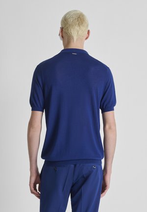 Osoba s kratkom plavom kosom nosi plavu majicu kratkih rukava i odgovarajuće plave hlače, stojeći okrenuta leđima prema kameri.