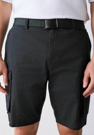 Czarne szorty cargo z bocznymi kieszeniami noszone z czarnym materiałowym paskiem i białą koszulą wpuszczoną do środka na stojącej osobie.