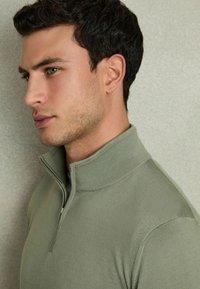 Pulover polo verde deschis cu fermoar, dotat cu guler ribat și o textură moale, prezentat pe un model cu un fundal neutru.