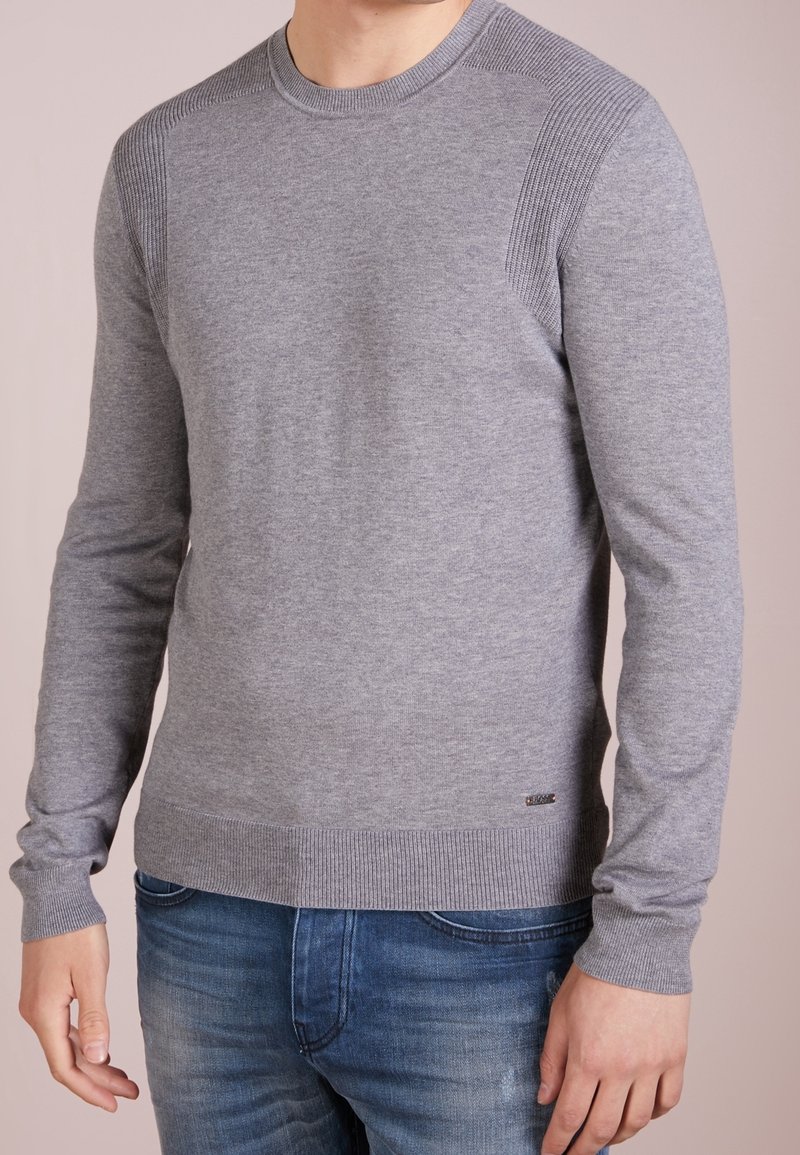 Grauer Langarmpullover mit gerippten Schulterakzenten und Bündchen. Verfügt über einen Rundhalsausschnitt und eine glatte Textur, kombiniert mit blauen Jeans.