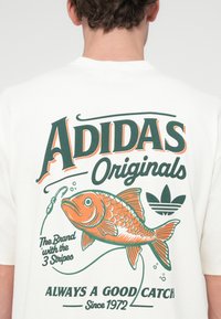 Λευκό κοντομάνικο μπλουζάκι με πορτοκαλί και πράσινο σχέδιο ψαριού, κείμενο "ADIDAS Originals" και λογότυπο με τρεις ρίγες.