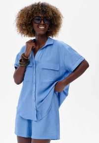 Light blue, short-sleeve button-up shirt with two front pockets, paired with matching shorts. Soft, textured fabric details enhance design.
