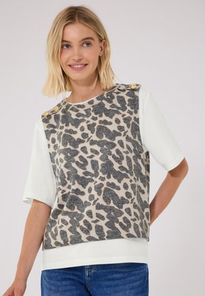 Giovane donna con capelli biondi che indossa una maglietta bianca a maniche corte sovrapposta a un gilet beige e nero con stampa animalier e bottoni dorati sulle spalle.