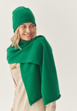 Zelená pletená čiapka a veľký pléd s hladkou textúrou, prehodený cez tričko neutrálnej farby. Dizajn je jednoduchý a moderný.