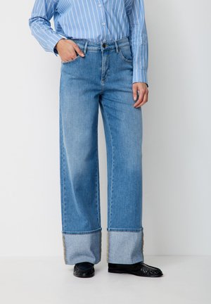 Person trägt hellblaue Jeans mit weitem Bein und umgeschlagenen Säumen, schwarze Leder-Loafer und ein blau gestreiftes Hemd mit Knöpfen.