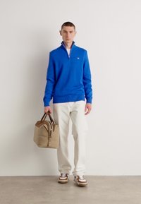 Blauer Strickpullover mit einem Reißverschlusskragen, weiße Hose, braun-weiße Sneakers und eine beige Tasche mit braunen Griffen. Einfaches und funktionales Outfit.