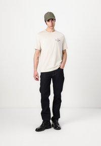 Beige cotton t-shirt with a small logo, dark blue denim jeans, and black leather shoes. Olive green cap completes the outfit.