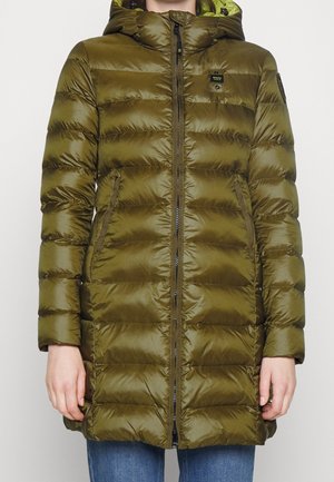 Veste longue matelassée vert olive avec capuche, fermeture éclair à l'avant, poches latérales et écusson noir avec logo sur la poitrine et la manche, portée avec un jean bleu.