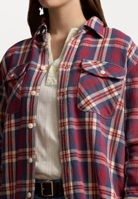 Frau trägt ein rot-blau-weiß kariertes Hemd mit Knöpfen über einem strukturierten weißen Henley-Oberteil, mit einem schwarzen Gürtel, der an der Taille sichtbar ist.