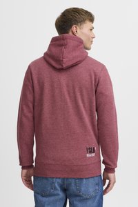 Czerwony sweter z kapturem i sznurkiem, o miękkiej teksturze i ribbrowanych mankietach; logo nadrukowane na dolnej części pleców. Noszony z dżinsami.